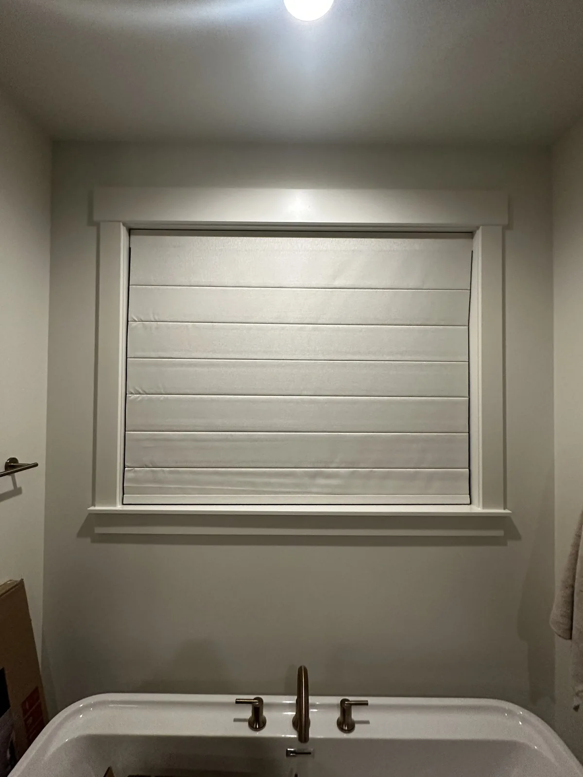 Light-colored Roman shade lowered over a bathroom window above a bathtub